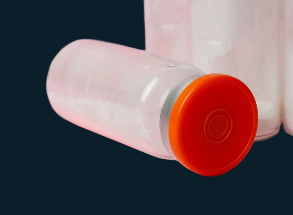 A glass vial of white lyophilized powder with an orange cap, lying on its side, cap toward the viewer